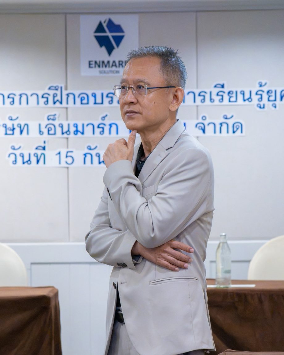 อ.ปกรณ์ วงศ์รัตนพิบูลย์ ยืนเอามือเท้าคางในห้องอบรมของ Enmark ครุ่นคิดเรื่องการประเมินก่อนพัฒนา