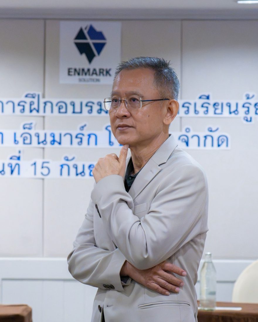 อ.ปกรณ์ วงศ์รัตนพิบูลย์ ยืนครุ่นคิดหน้าผนังโลโก้ Enmark ชวนองค์กรส่งบุคลากรเข้าโปรแกรมพัฒนาที่เชื่อมกับเป้าหมายธุรกิจ