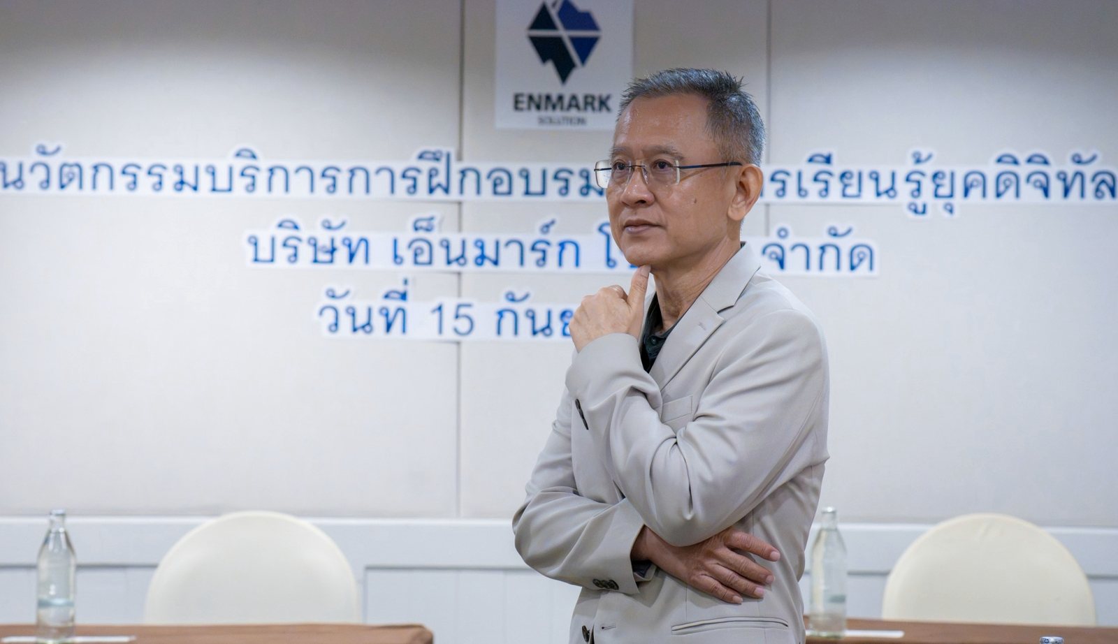อ.ปกรณ์ วงศ์รัตนพิบูลย์ ยืนครุ่นคิดหน้าผนังโลโก้ Enmark ผู้วางแนวทางมาตรฐานการพัฒนาบุคลากรของสถาบัน