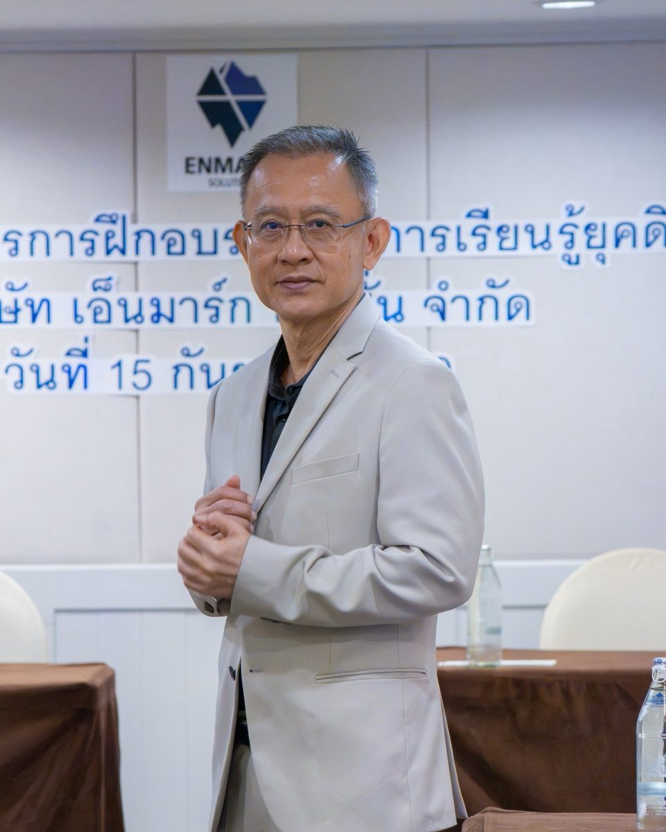 อ.ปกรณ์ วงศ์รัตนพิบูลย์ ยืนหน้าผนังโลโก้ Enmark ผู้วางแนวทางมาตรฐานการเชื่อมการพัฒนาบุคลากรกับเป้าหมายธุรกิจ