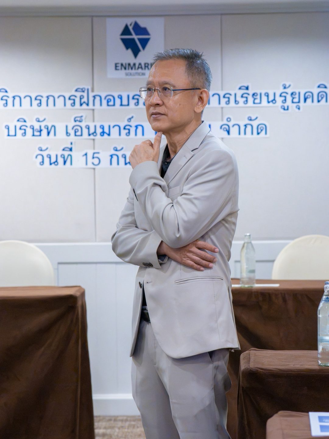 อ.ปกรณ์ วงศ์รัตนพิบูลย์ ยืนเท้าคางครุ่นคิด หน้าผนังโลโก้ Enmark Solution