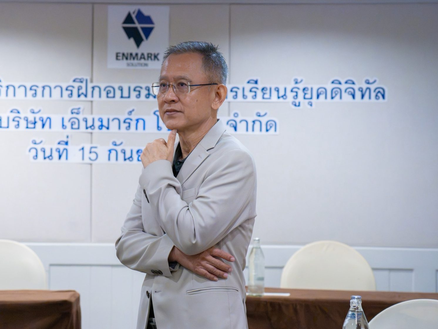 อ.ปกรณ์ วงศ์รัตนพิบูลย์ ยืนเอามือแตะคาง ครุ่นคิดถึงโจทย์ธุรกิจ หน้าผนังโลโก้ Enmark Solution
