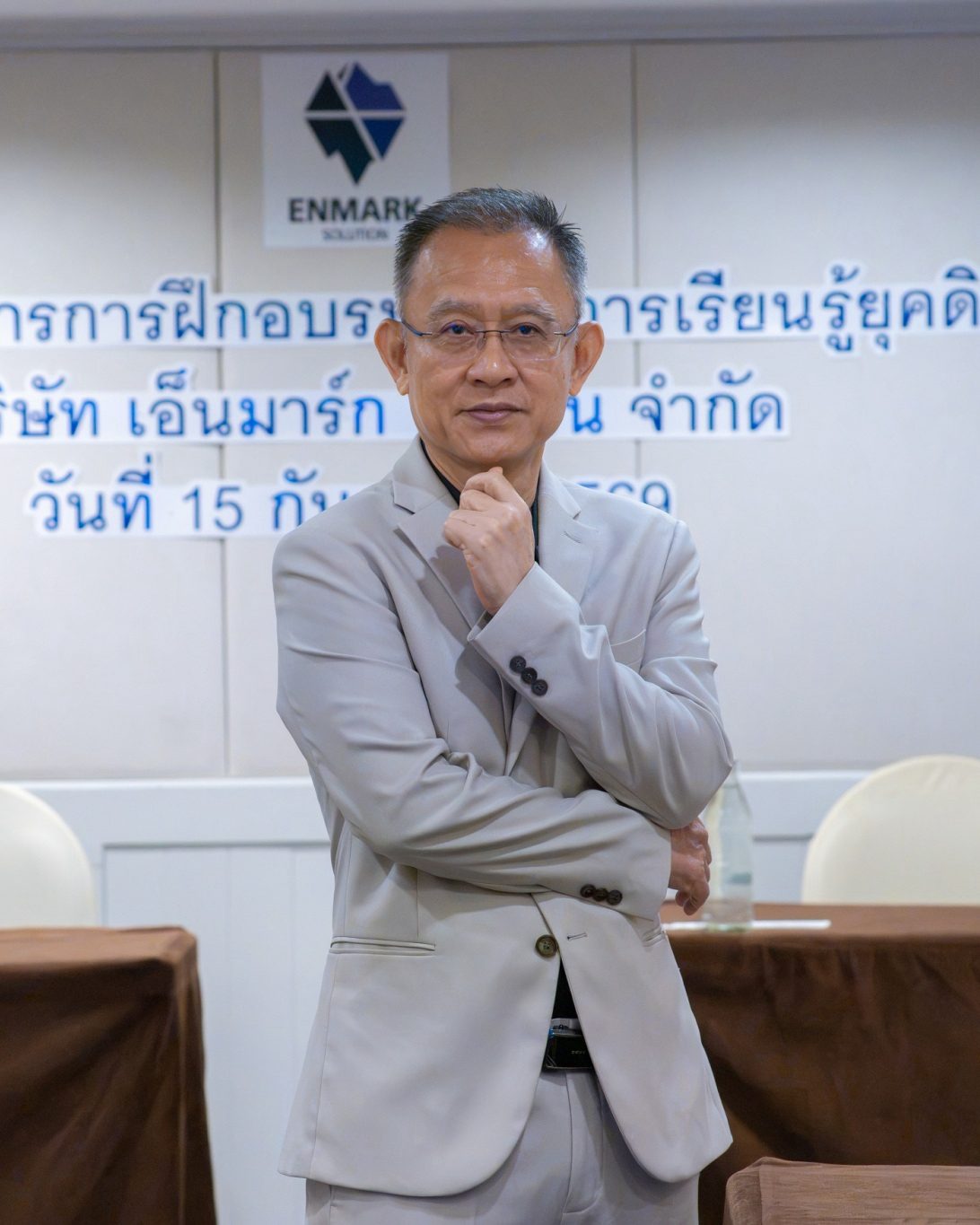 อ.ปกรณ์ วงศ์รัตนพิบูลย์ ยืนกอดอกเอามือแตะคาง หน้าผนังโลโก้ Enmark Solution